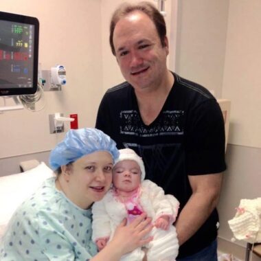 A woman wearing a hospital gown and bouffant cap sits in a hospital bed holding her 4-month-old daughter. Her husband stands behind them, helping to hold up the baby. Both parents are smiling at the camera while the baby's eyes are closed.