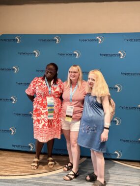 Three women pose with their arms around each other in front of a wall. 