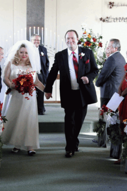 A bride and groom hold hands while walking down the aisle of a church. The bride is carrying a red bouquet of flowers. A pastor looks on from behind the couple.