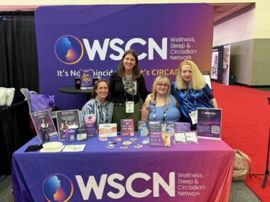 Four women sit behind a table covered with information and products. Purple banners behind and in front of them say 