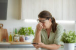 A woman holds a hand to her face while sitting at a kitchen counter looking at a phone.