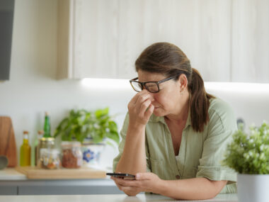 Woman with glasses looking tired while holding a phone in a kitchen.