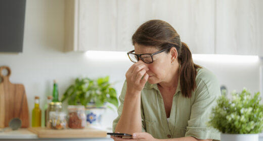 Woman with glasses looking tired while holding a phone in a kitchen.