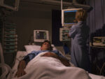 A man sleeps in a hospital bed while a healthcare professional monitors medical equipment and vital-sign displays.