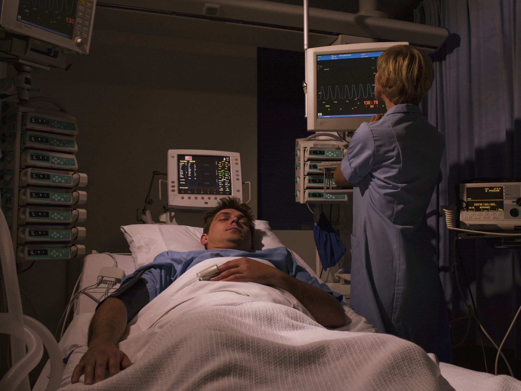 A man sleeps in a hospital bed while a healthcare professional monitors medical equipment and vital-sign displays.