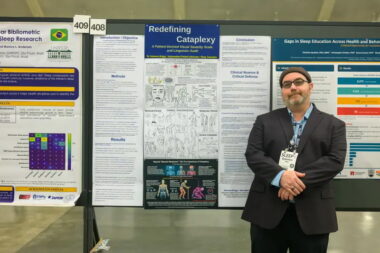 A man in dress clothes poses with his arms crossed in front of him, in front of a large scientific poster hanging on a wall. 