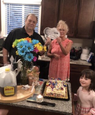A grandmother and a grandfather stand behind a kitchen counter as their granddaughter stands off to the side. There are brightly colored flowers and cake on the counter, and the adults are holding up a World Narcolepsy Day sign.