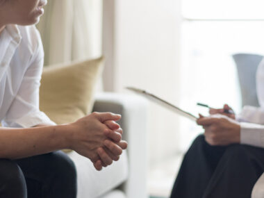 A person sits across from a healthcare professional holding a clipboard during a consultation.
