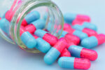 Half-pink, half-blue capsules are seen spilling out of a glass bottle lying on its side.