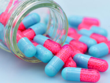 Half-pink, half-blue capsules are seen spilling out of a glass bottle lying on its side.