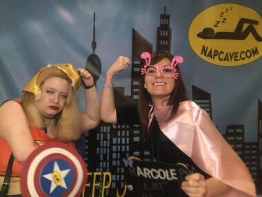 Two women dressed as superhero figures strike a "muscle" pose in a room designed as a comic book city. A yellow sign behind them identifies it as the "Nap Cave," with a stick-figure logo of a person lying on their back with "ZZZZZZ" above it. The posing is meant to be both silly and empowering.