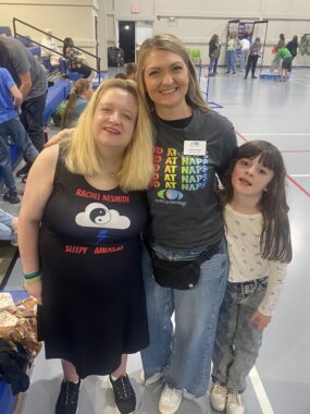 Two women and one child smile for a photo. They're standing next to bleachers overlooking an indoor court.