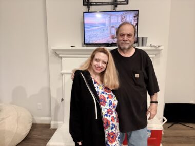 A middle-age woman stands next to her father in what appears to be a living room. The man has his arm around his daughter's shoulder, and both are smiling at the camera.