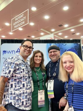 Two men and two women gather for a photo in front of the PWN4PWN booth at a convention. They're all wearing lanyards and smiling at the camera.