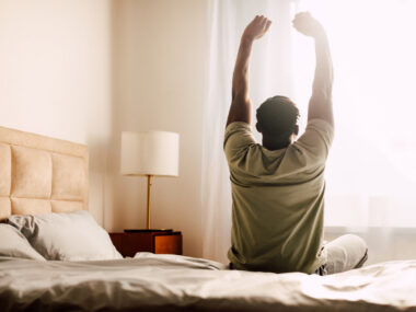 A person is sitting on a bed is seen from behind stretching as light comes in a curtained window.