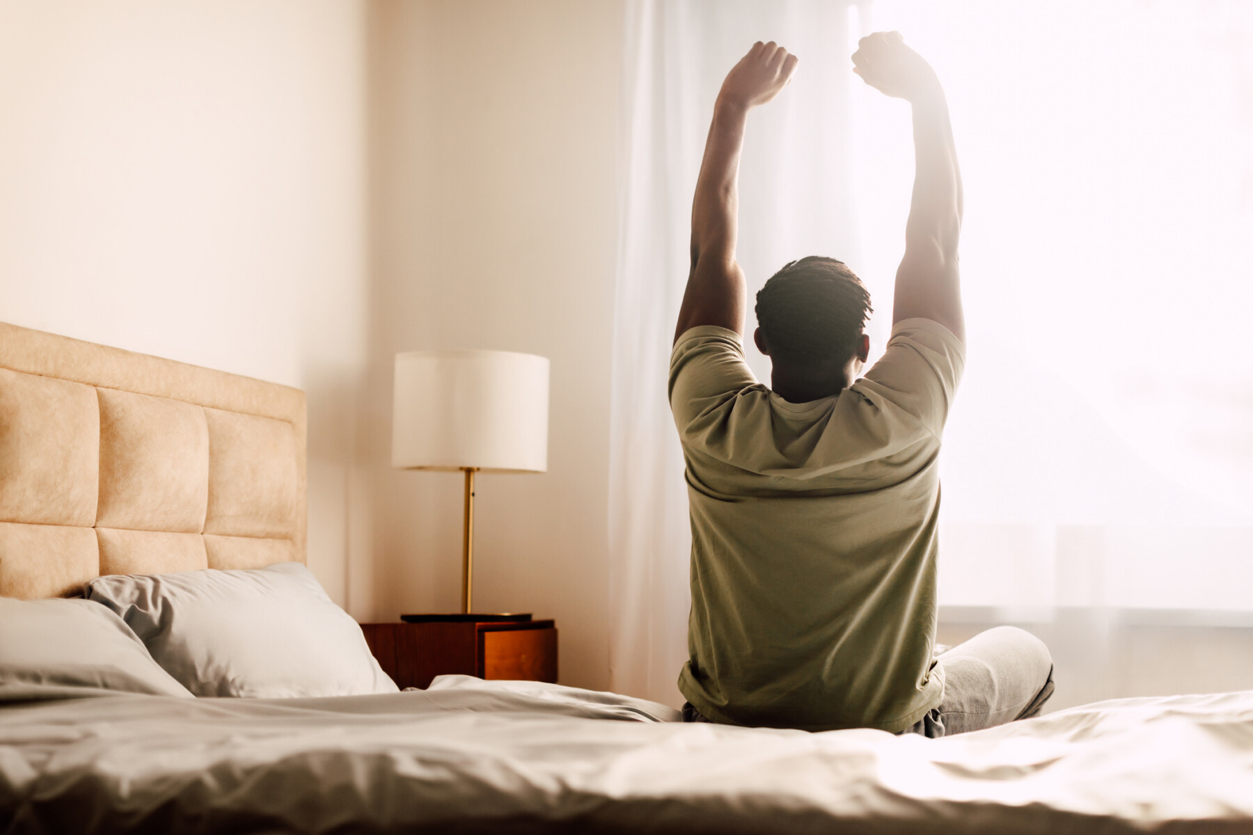 A person is sitting on a bed is seen from behind stretching as light comes in a curtained window.