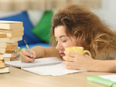 A tired-looking girl sits at a desk writing on a paper, with a mug in one hand.