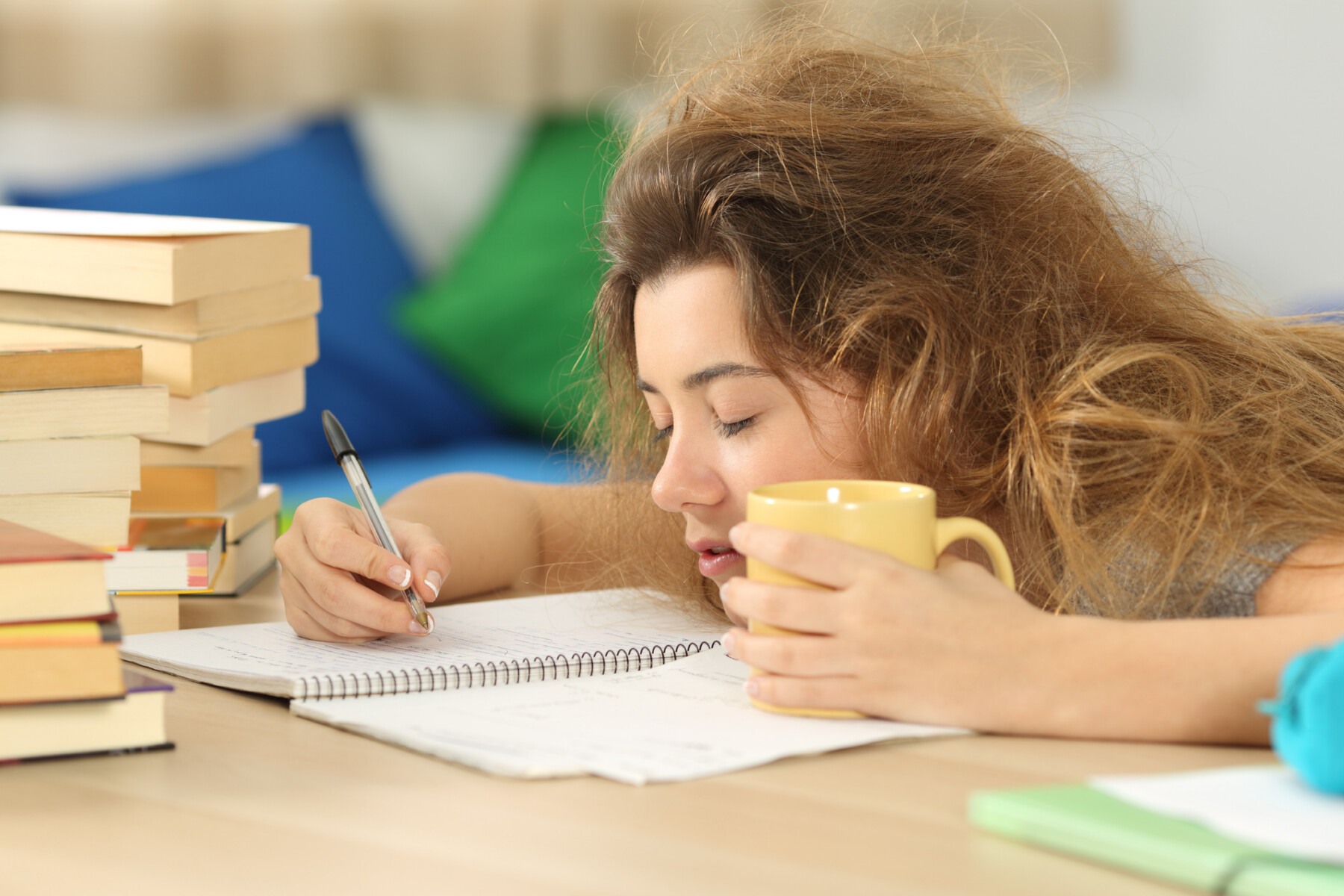 A tired-looking girl sits at a desk writing on a paper, with a mug in one hand.