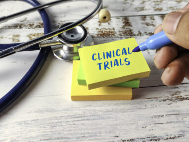 A hand holding a blue marker writes “Clinical Trials” on a yellow sticky note beside a stethoscope.