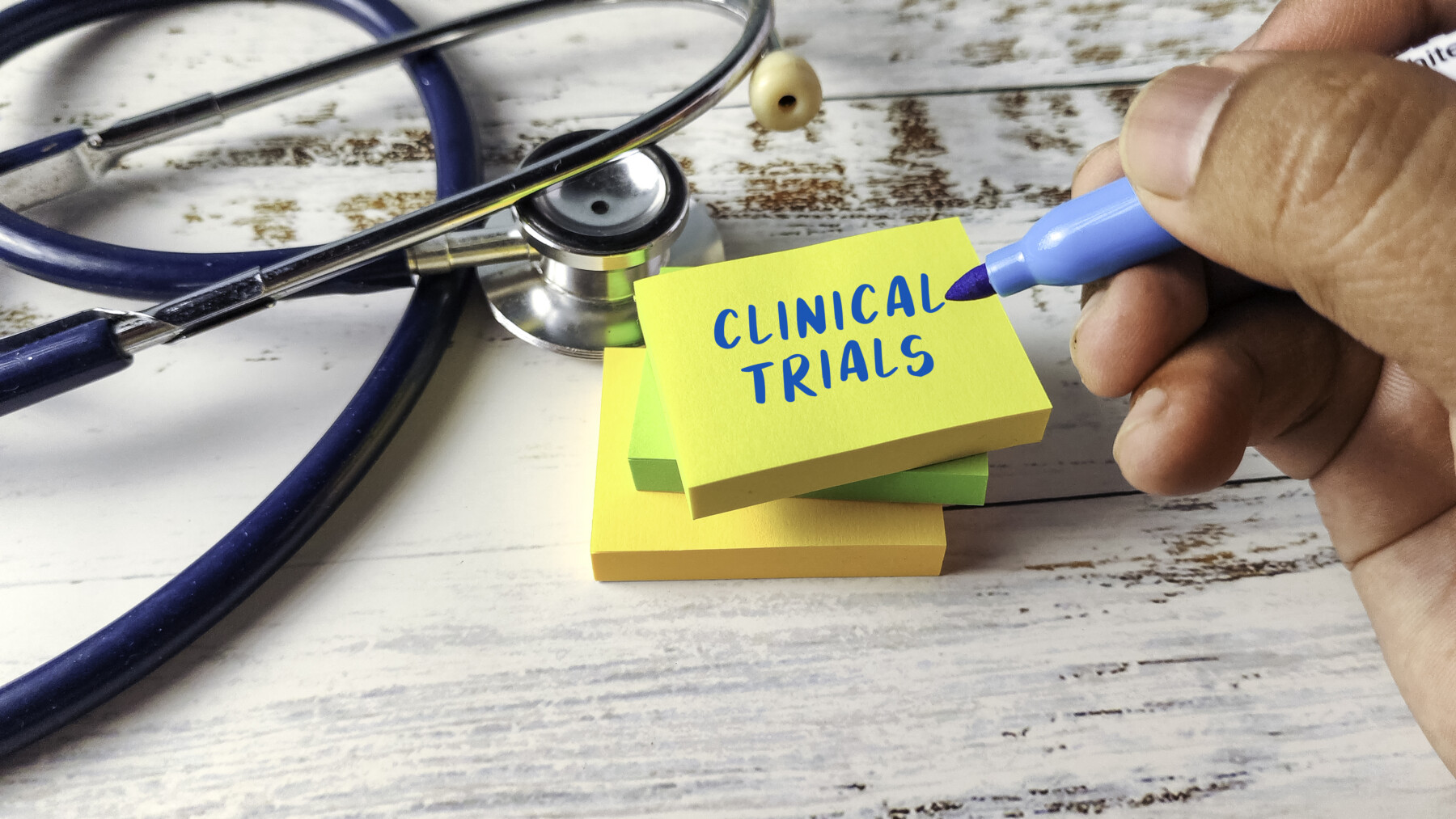A hand holding a blue marker writes “Clinical Trials” on a yellow sticky note beside a stethoscope.